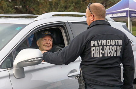 Photo of a person in a vehicle speaking with a person wearing a "Plymouth Fire-Rescue" shirt at a drive-thru event